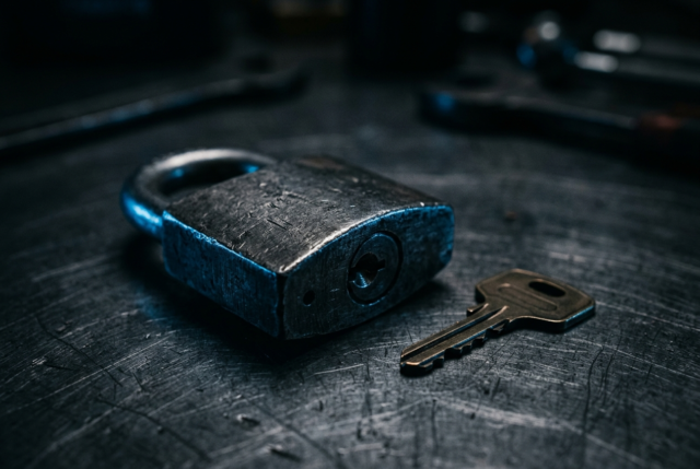 A heavy steel padlock and key resting on a dark brushed metal surface in cool light A heavy steel padlock and key resting on a dark brushed metal surface in cool light